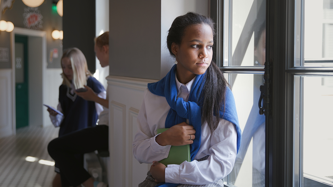 Portrait of sad African-American teen student sitting on window sill in corridor. Upset schoolgirl having problems with education or failing examination looking out of window