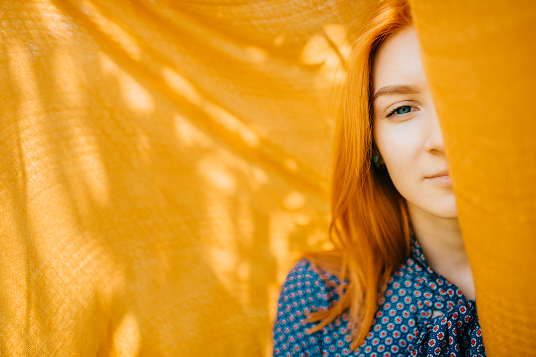 Beautiful girl with red hair hiding half face behind orange blanket with abstract shadows on background. Mystery psychological woman portrait. Young pensive teen looking out of cover. Alter ego.