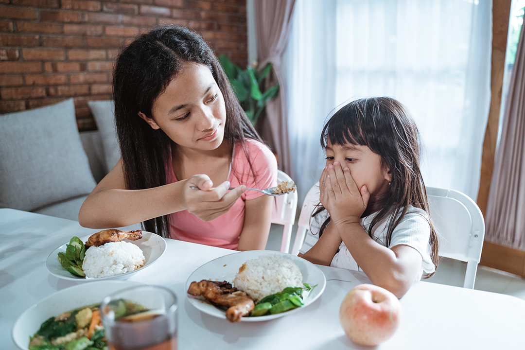 little girl refuses to eat and her older sister is annoyed when eating together at the dining table against the dining room background