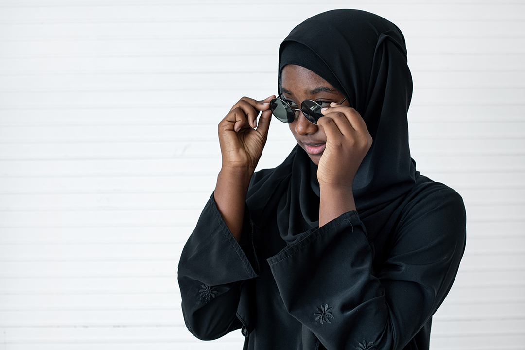African Muslim pre-teen wearing black hijab and wearing sun glasses, on white wall background
