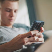 Caucasian teenager chats in messaging app with friends or watches content using mobile phone while sitting on sofa in living room. Teen boy spending weekend at home. Lifestyle concept. Close Up.