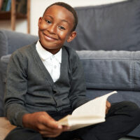 Cute brilliant smart child of african american ethnicity study at home, doing homework, sitting on floor with book in hands smiling at camera with enthusiasm, happy to get new knowledge