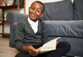 Cute brilliant smart child of african american ethnicity study at home, doing homework, sitting on floor with book in hands smiling at camera with enthusiasm, happy to get new knowledge