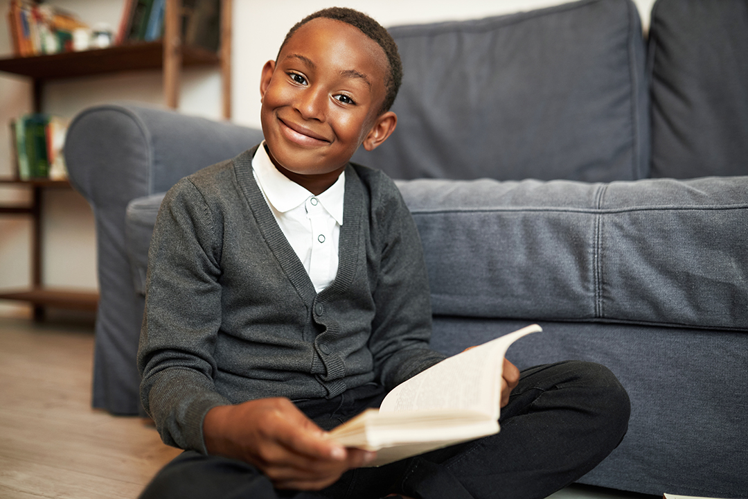 Cute brilliant smart child of african american ethnicity study at home, doing homework, sitting on floor with book in hands smiling at camera with enthusiasm, happy to get new knowledge