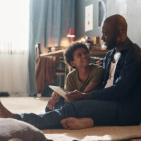 Full length portrait of African American single father and smiling son using tablet and chatting sitting on floor in harsh sunlight copy space