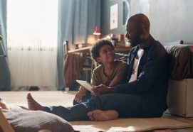 Full length portrait of African American single father and smiling son using tablet and chatting sitting on floor in harsh sunlight copy space
