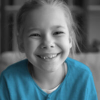 Close up headshot portrait of cute happy small preschooler girl with toothy smile look at camera posing, funny little child sit relax have fun on sofa in living room, feel optimistic and positive