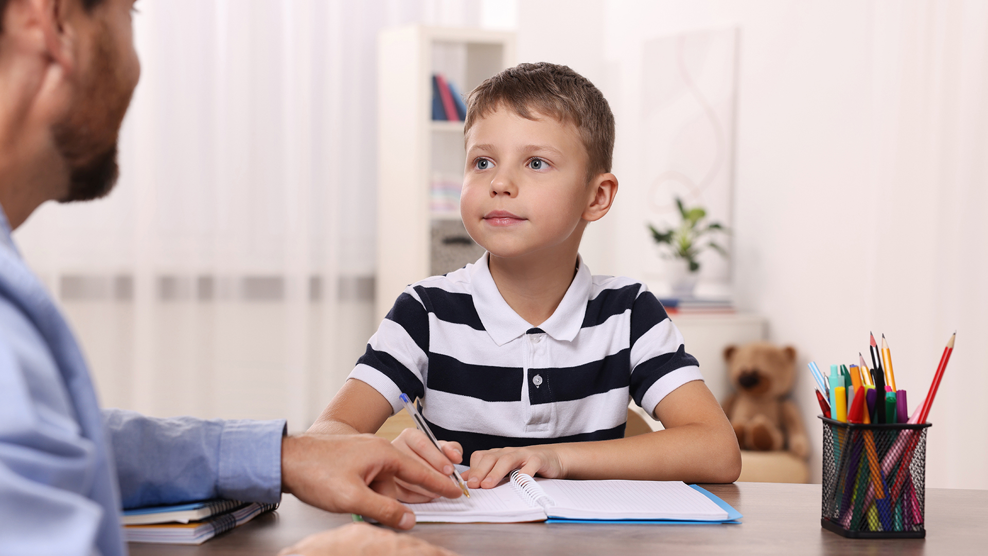 young boy writing at desk with adult supervision