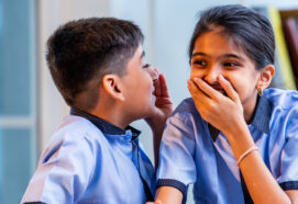 Indian School Kids Sharing Gossip with Friend in a Classroom While Wearing School Uniforms, Whispering Secret Conversations and Laughing Quietly During Leisure Moment Between Study Sessions in school