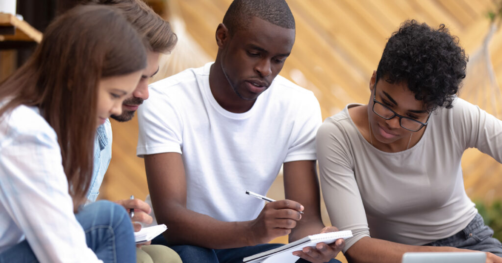 Beautiful smart girls handsome clever guys sitting in cozy place studying writing notes thoughts in notepad. Skilled man helps to teammates understand lecture learn preparing for examination together