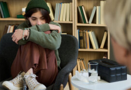 Teenage girl sitting curled up in chair hugging knees during group therapy session, looking down with thoughtful expression while adult woman listening nearby