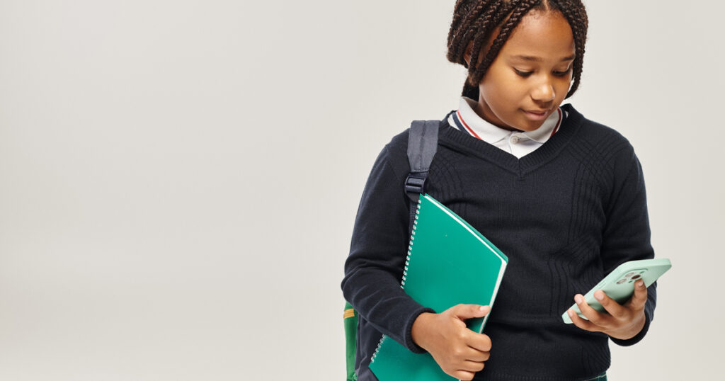 preteen african american schoolgirl in uniform using smartphone and holding notebook on grey