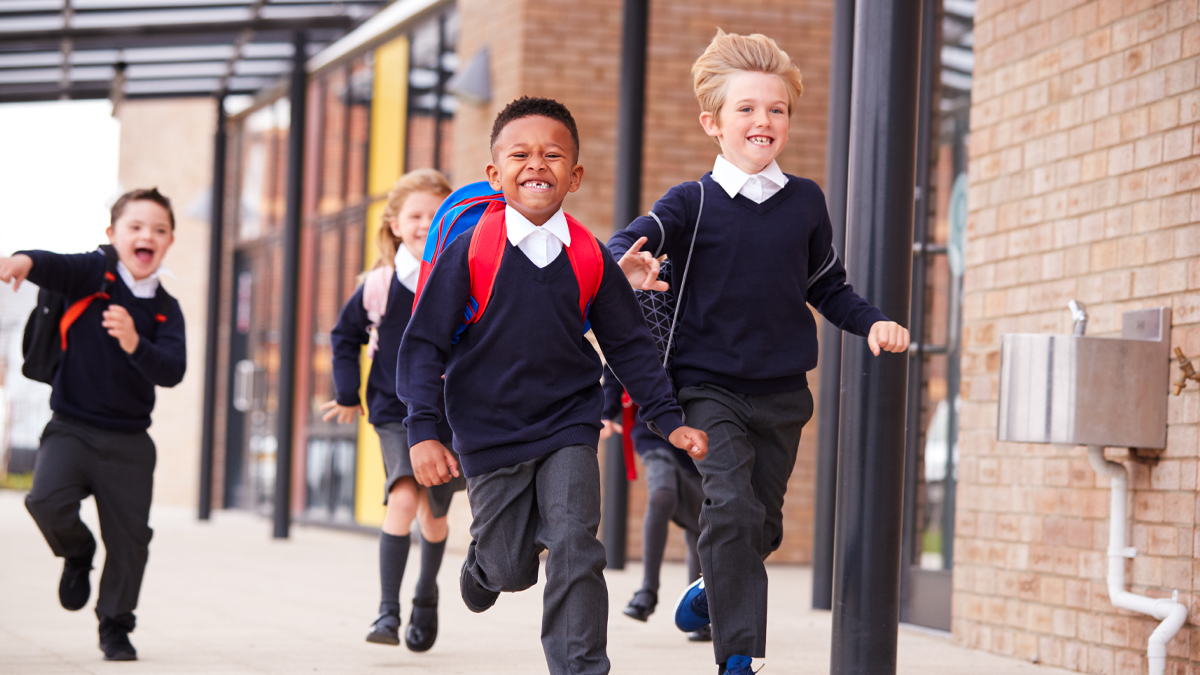 Happy primary school kids, wearing school uniforms and backpacks, running on a walkway outside their school building, front view, close up