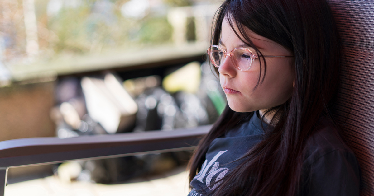 Sad dark-haired girl with glasses sitting at a table in the backyard. Cute children's difficulties concept