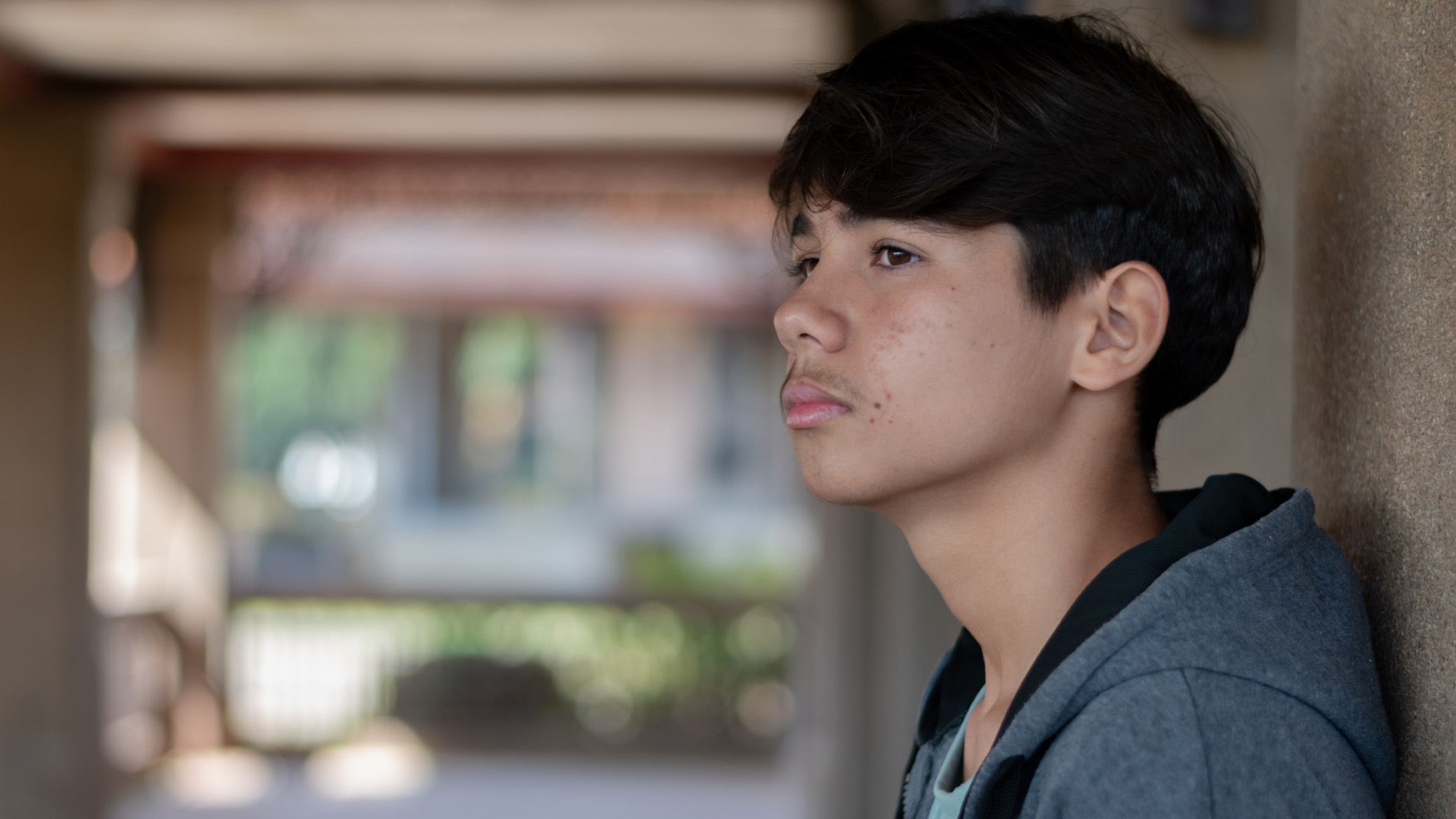 Pensive side portrait of a teen boy in a grey hoodie, leaning against a pillar in an outdoor walkway Pensive side portrait of a teen boy in a grey hoodie, leaning against a pillar in an outdoor walkway