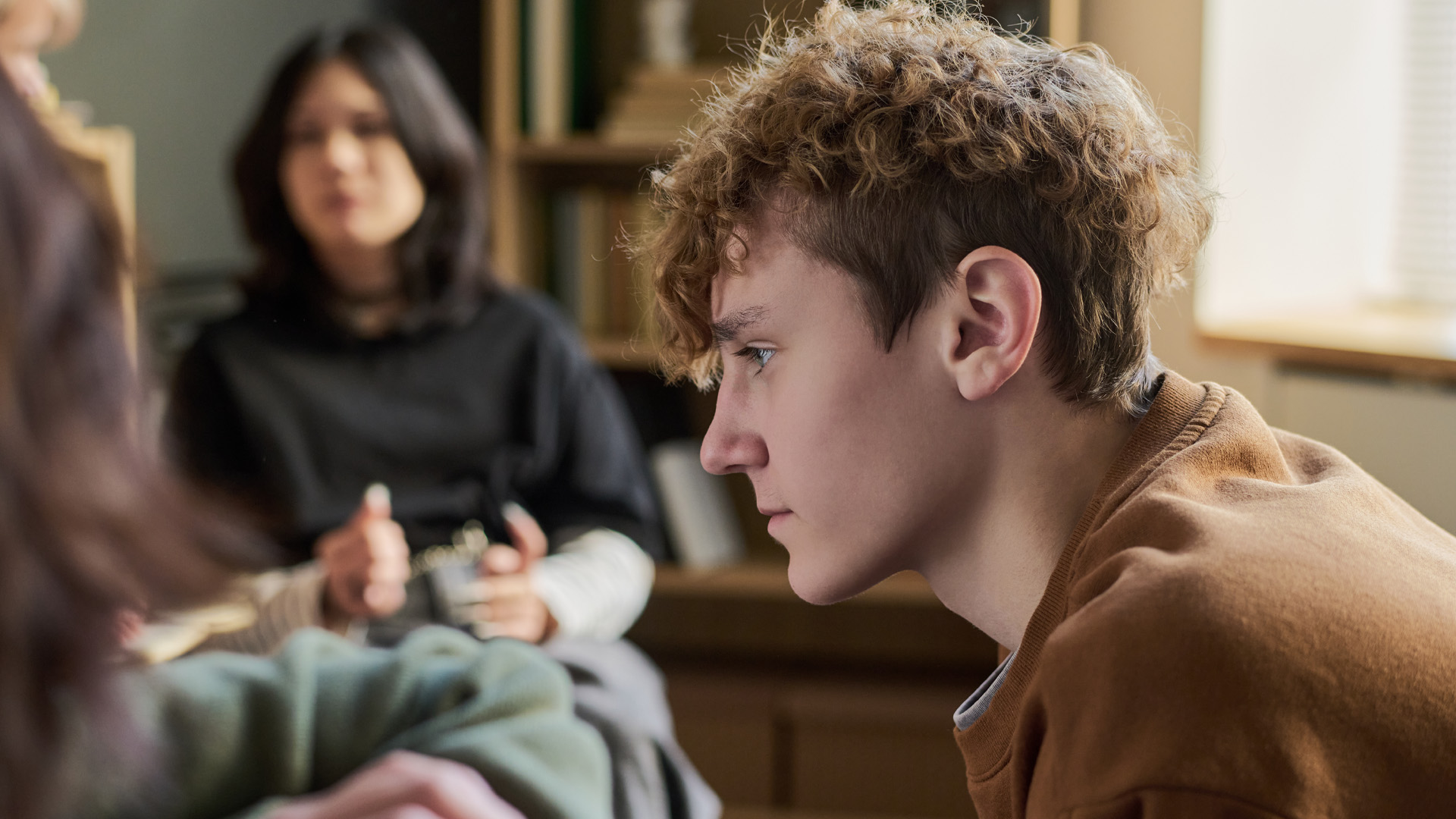 Teenager boy sitting in profile listening attentively within a group with multiethnic young people and teenagers in background participating in discussion Teenager boy sitting in profile listening attentively within a group with multiethnic young people and teenagers in background participating in discussion
