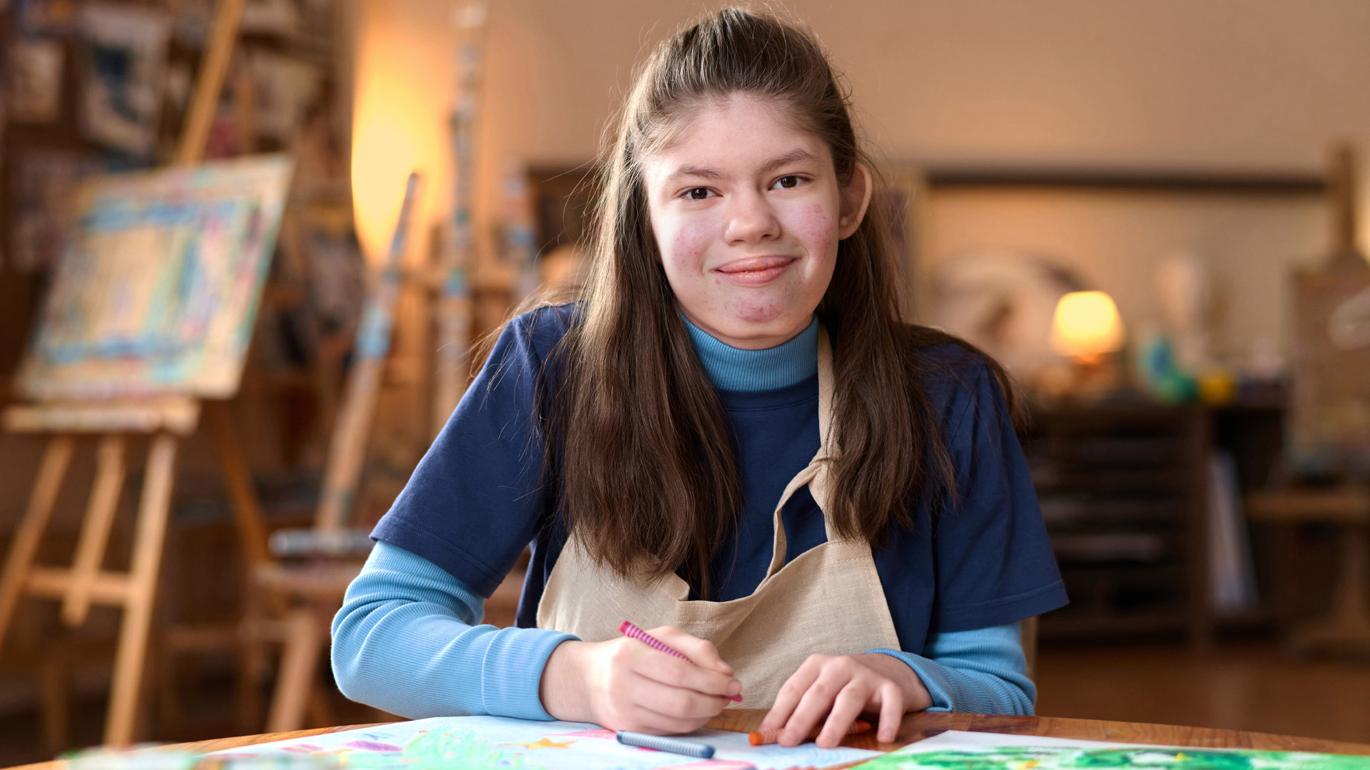 Front view portrait of young girl with Williams syndrome smiling at camera sitting at table in art studio