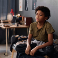 Full length portrait of teenage boy sitting on bed in his room and looking away