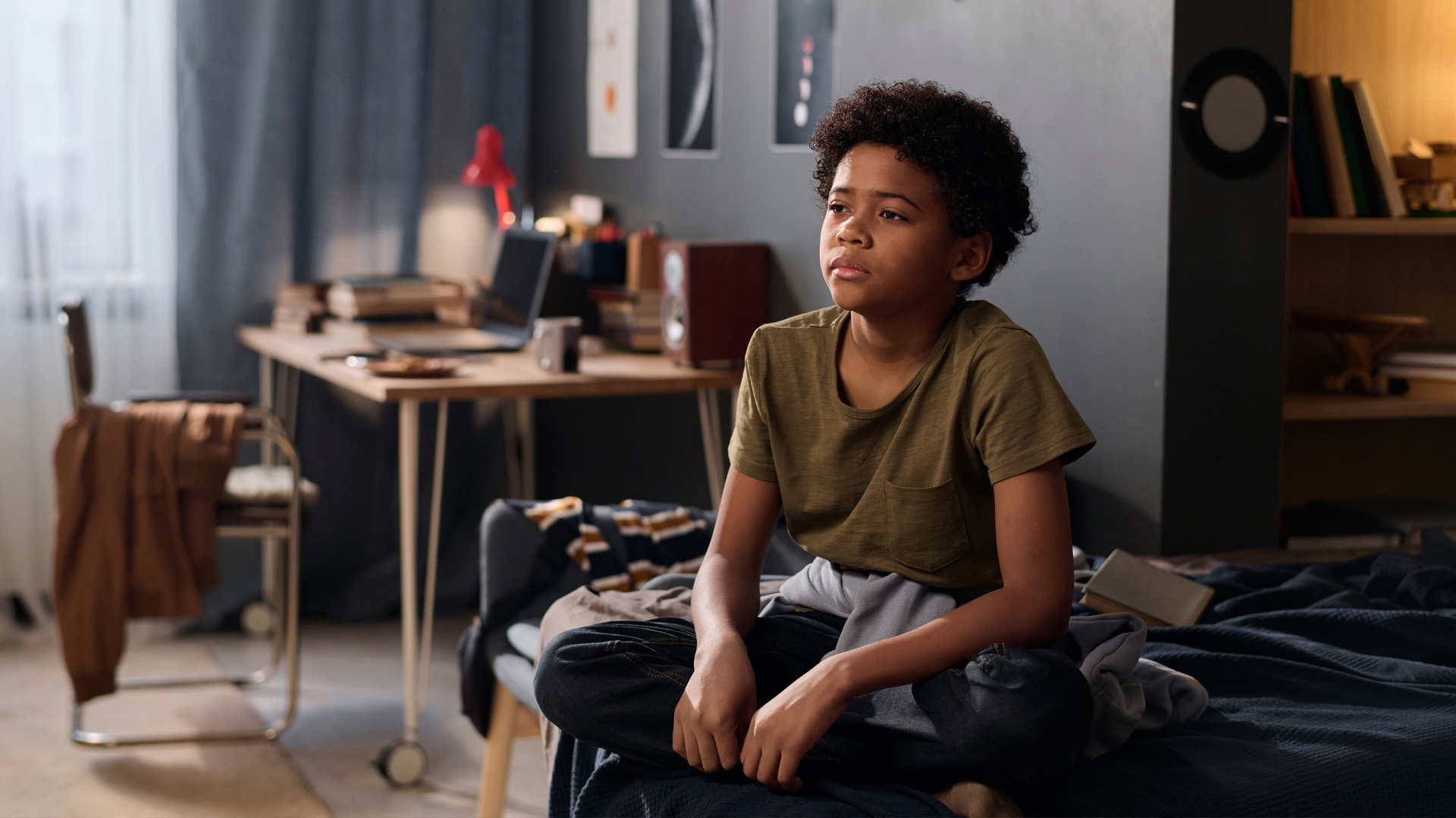 Full length portrait of teenage boy sitting on bed in his room and looking away