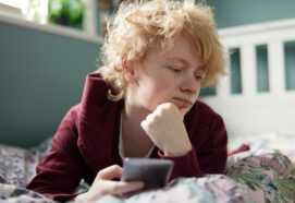 Unhappy Teenage Girl Lying On Bed At Home With Mobile Phone
